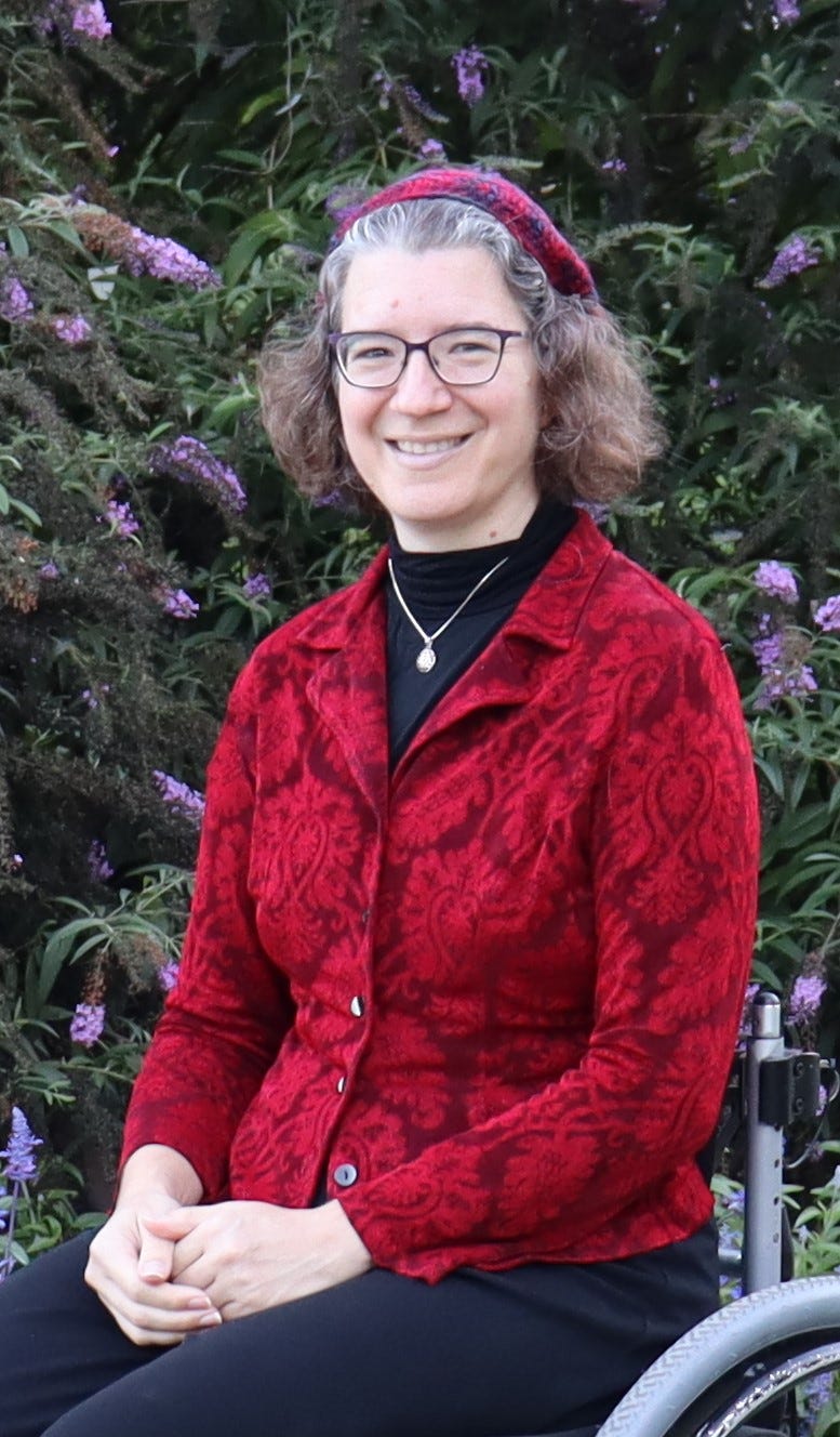 Julia Watts Belser, a white Jewish woman with curly brown and silvered hair, sits happily in her wheelchair in front of a flowering bush. She's wearing a patterned red blazer and red kippah (beret) to match.