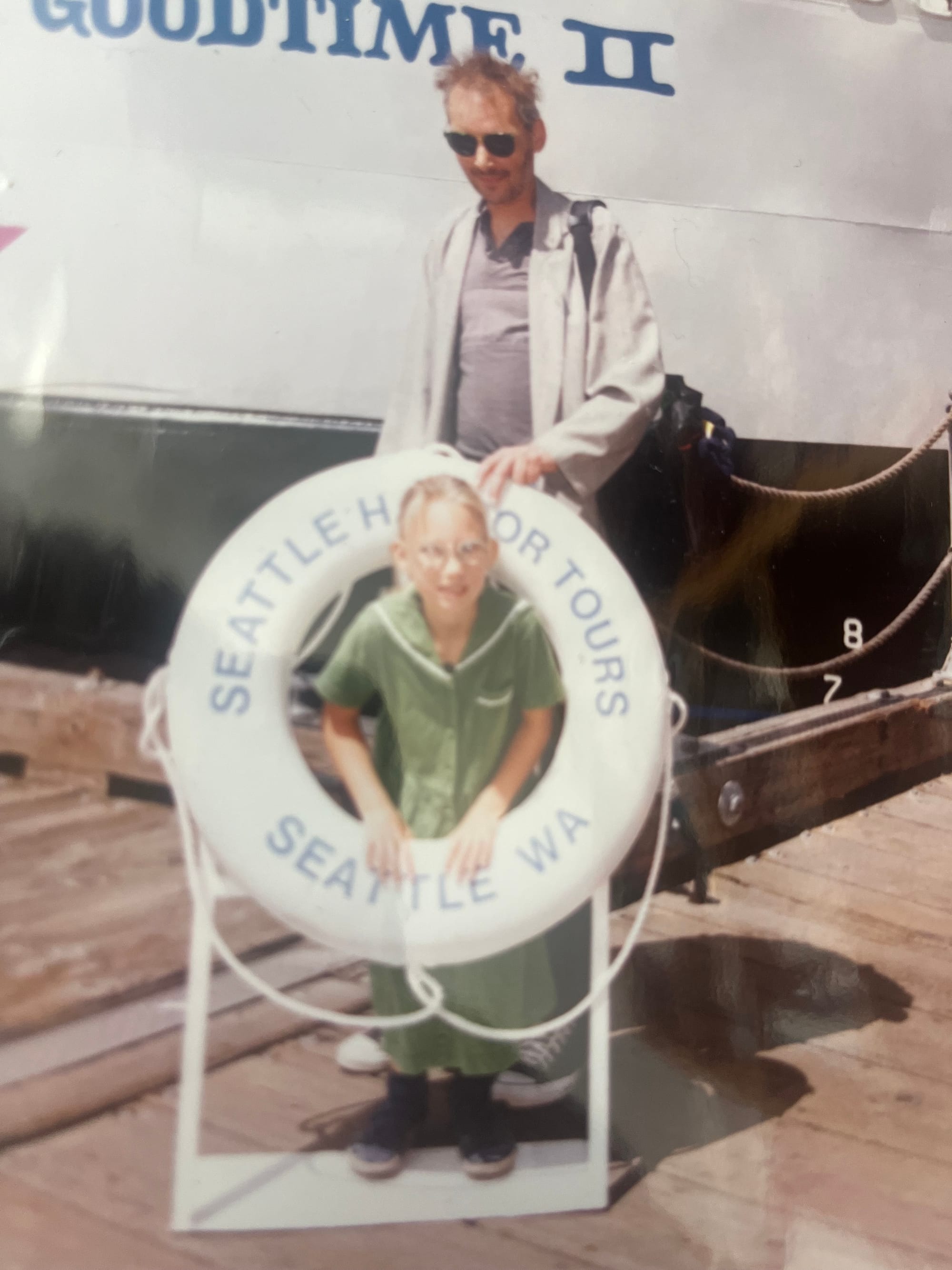 White man in sunglasses near a boat looks down fondly at small white blond child in glasses and a green dress sticking her head through a lifesaver on which is written, "Seattle Harbor Tours, Seattle, WA." 